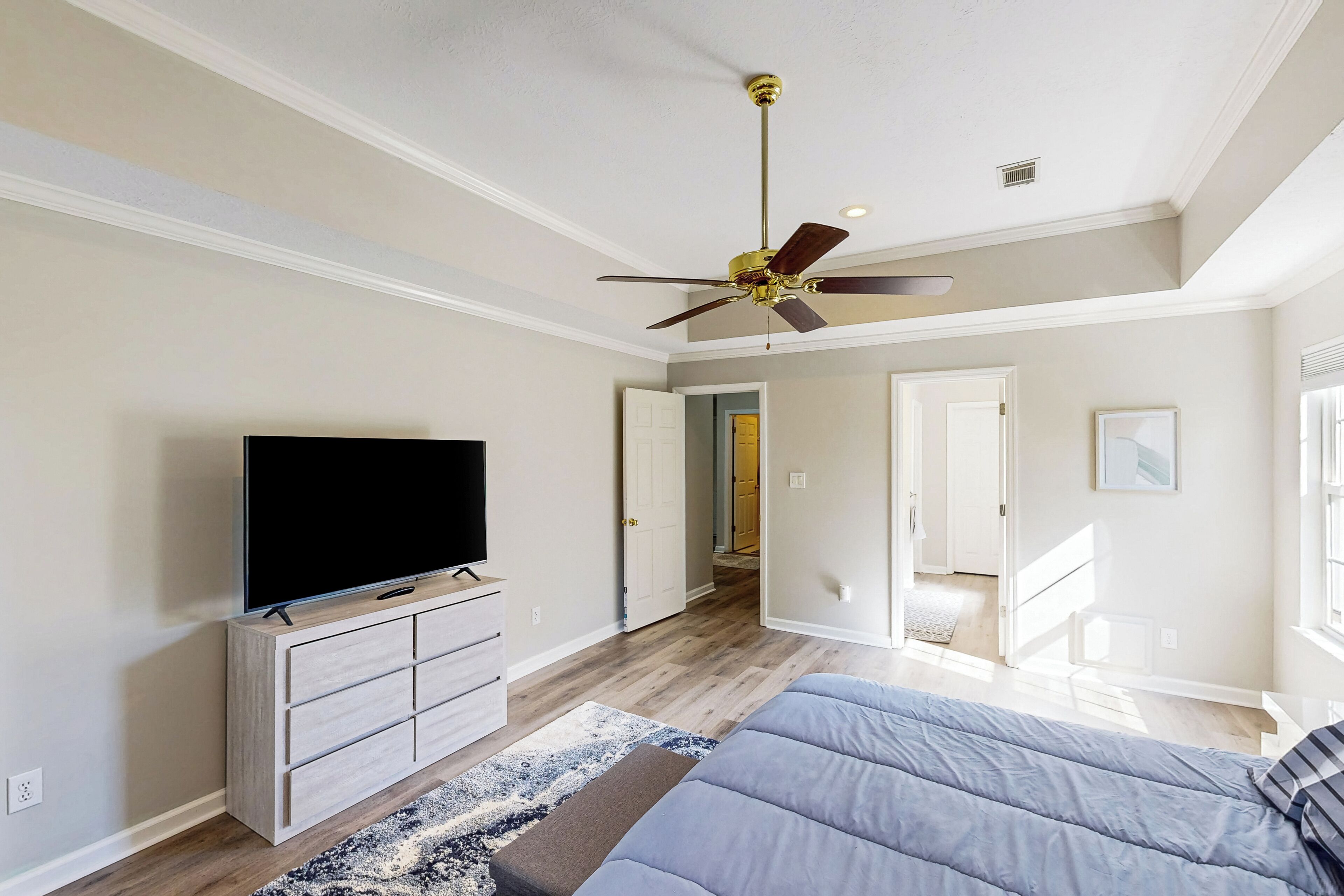 Une chambre à coucher avec un lit, un téléviseur posé sur une commode, un ventilateur de plafond et un couloir menant à une autre pièce.