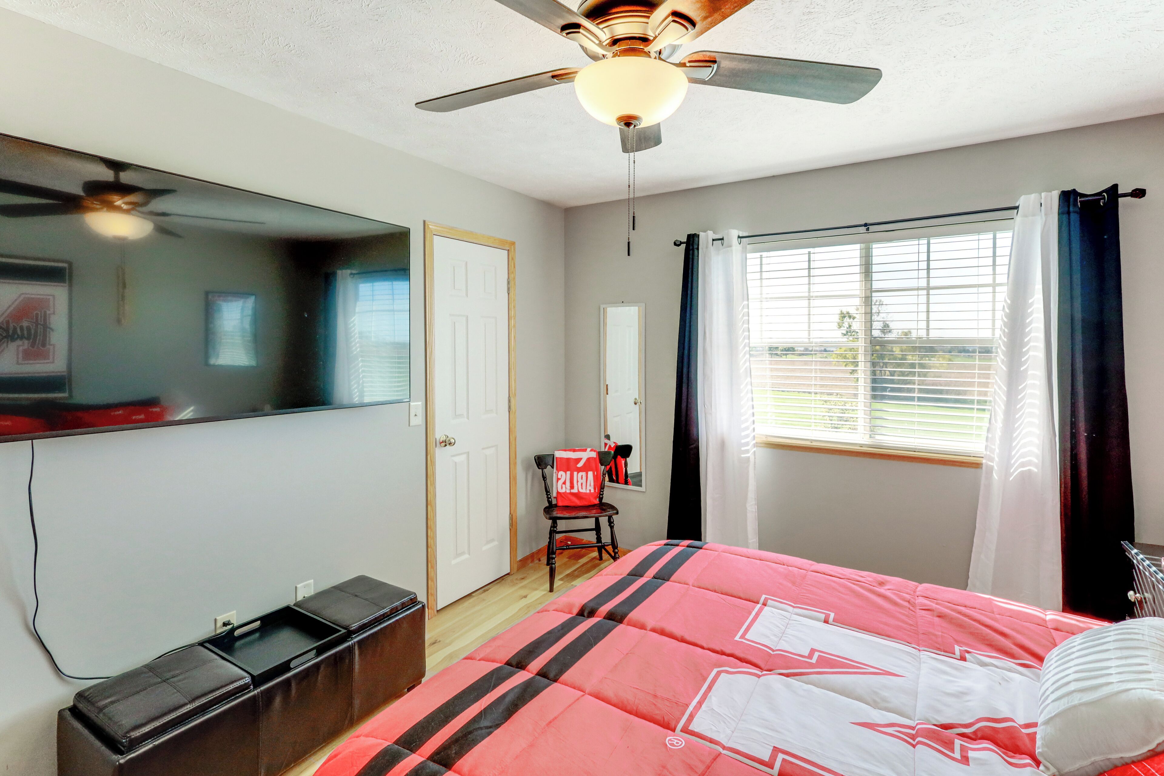 A bedroom with a bed, ceiling fan, window with curtains, and a chair.