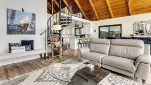 Un vaste espace de vie avec un plafond haut, un canapé, une table basse en verre, un tapis à motifs, un fauteuil, un escalier et une cuisine à l’arrière-plan.