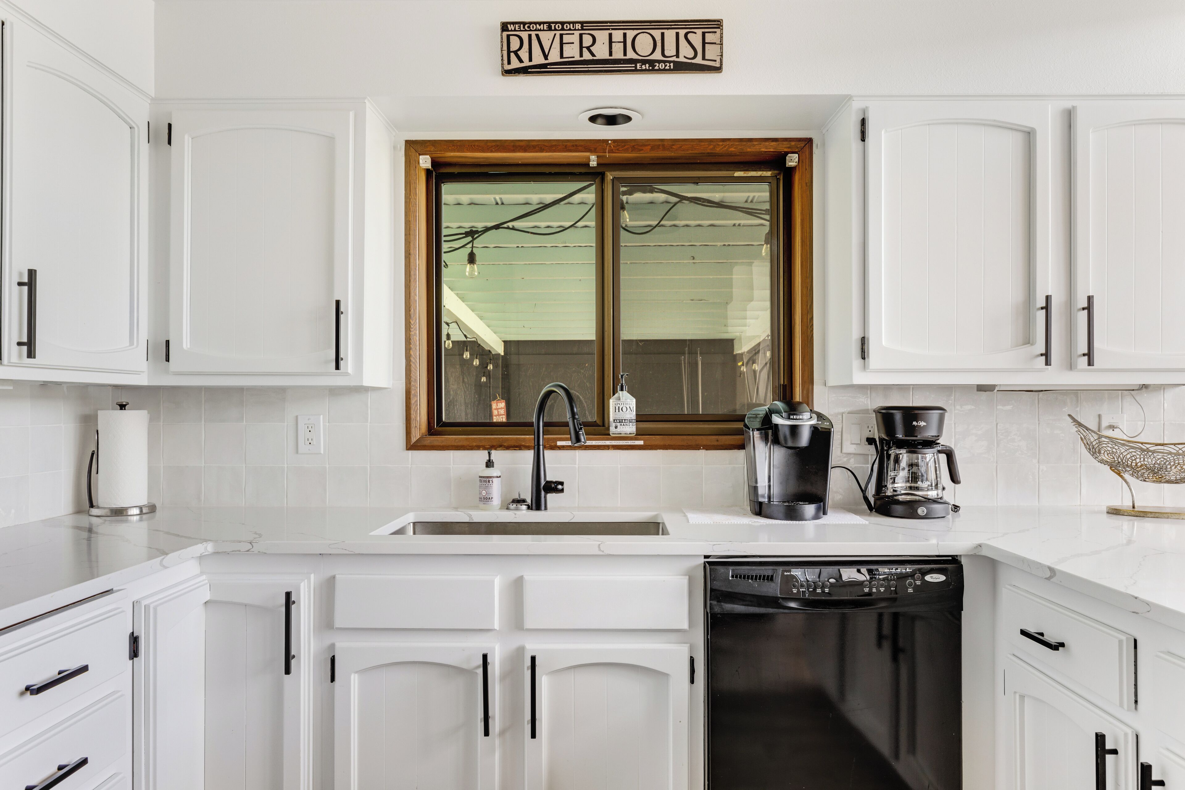 A modern kitchen with white cabinetry, a built-in dishwasher, a coffee maker, and a window with a wooden frame.