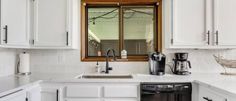 A modern kitchen with white cabinetry, a built-in dishwasher, a coffee maker, and a window with a wooden frame.