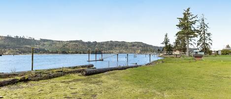 Un área cubierta de césped con un muelle de madera que se adentra en un lago, rodeado de colinas y árboles.
