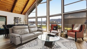 Un salon spacieux avec un plafond haut, de grandes fenêtres, un canapé en cuir, une table basse en verre et une vue sur un lac et des montagnes.