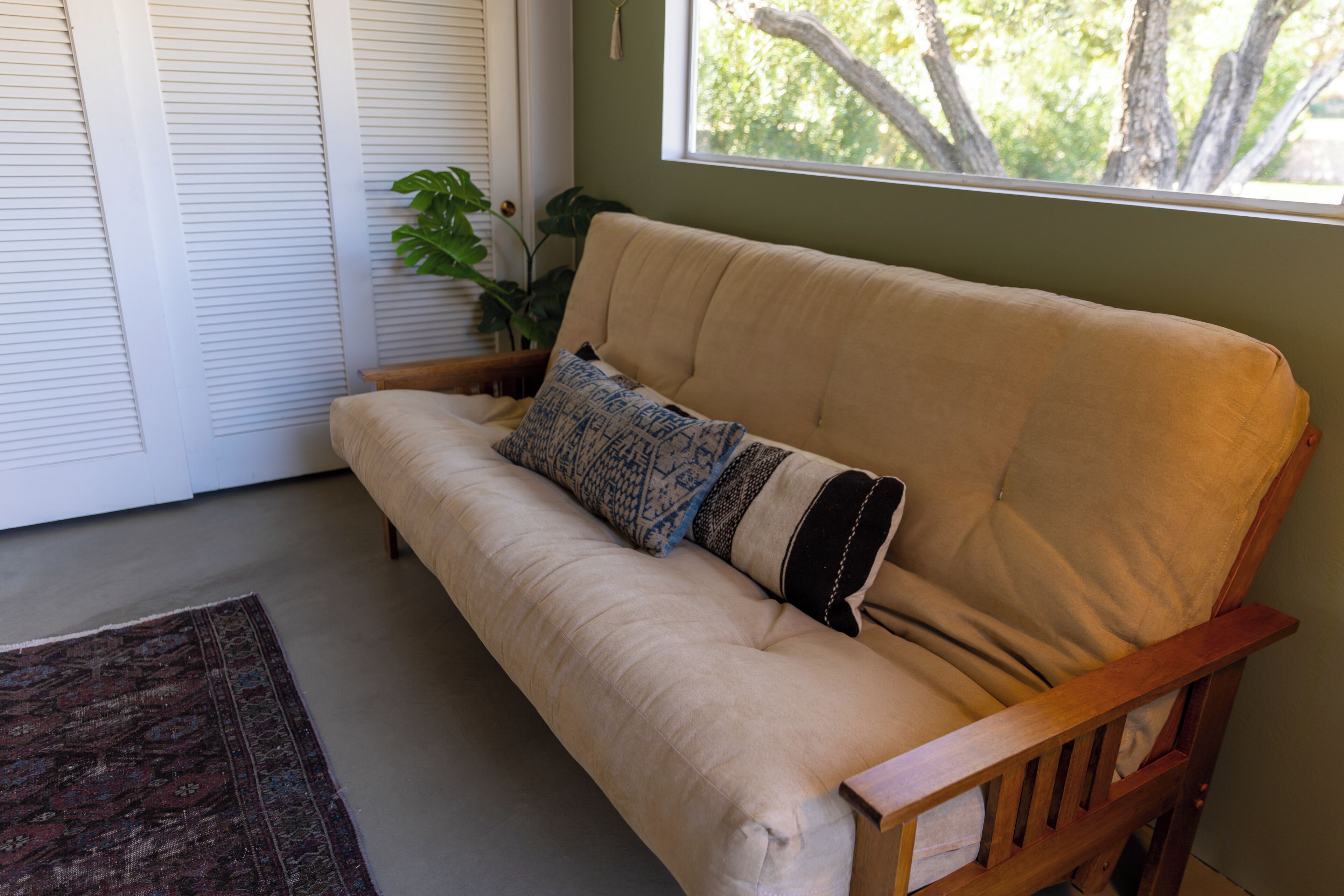 A beige sofa with a patterned pillow, a wooden armrest, and a window with a view of trees.
