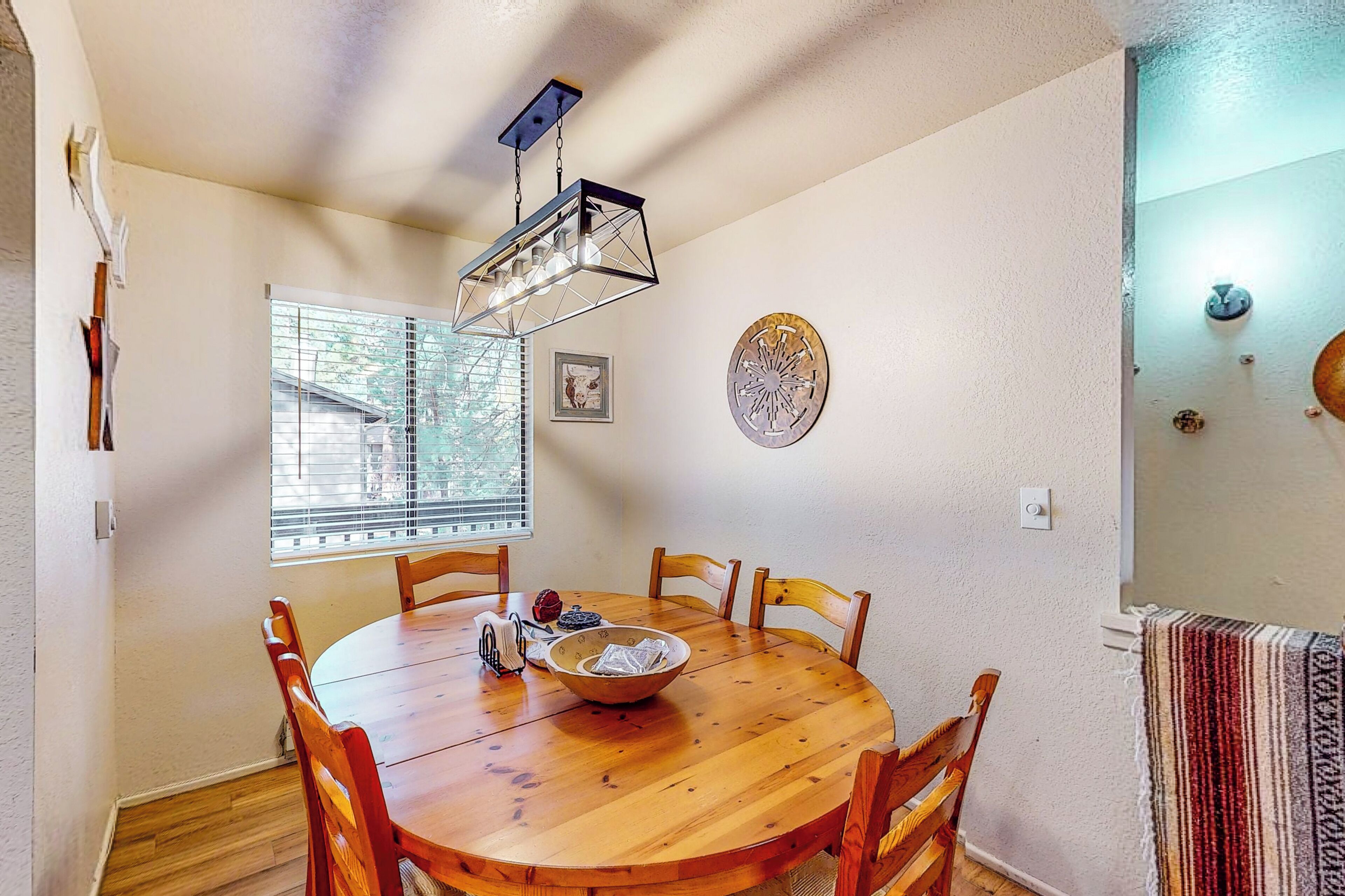 Un coin repas comprenant une table en bois, six chaises, un luminaire suspendu, une fenêtre avec des stores et une horloge fixée au mur.