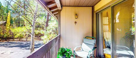 A balcony with a wooden railing, a glass table with a potted plant, a green chair, and a sliding glass door.