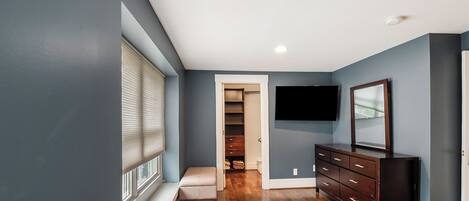 Una habitación con piso de madera, un aparador oscuro, un televisor de pantalla plana montado en la pared y una ventana con persianas.