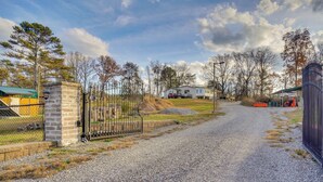 A gravel driveway leading to a gated entrance with a stone pillar, a house in the background, and a fenced area with trees and a parked car. - Pet-friendly Apt Near Ocoee River & Waterfalls (Etowah)