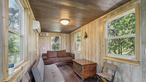 A cabin with a wooden interior, a brown sofa, a wooden chest, and a window overlooking greenery. - Fishing Pond & Treehouse: Tranquil Maysville Cabin (Maysville)