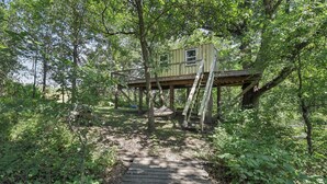 A treehouse elevated on stilts in a forested area with a wooden pathway leading to it. - Fishing Pond & Treehouse: Tranquil Maysville Cabin (Maysville)