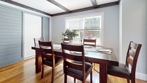 Un comedor con una mesa de madera dispuesta para cuatro, flanqueada por sillas de madera, una ventana con persianas y un techo con vigas a la vista.
