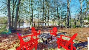A campsite with four red Adirondack chairs and a table, surrounded by trees and a body of water. - 3BR Riverfront Home w/ Kayaks & Game Room (Seaford)