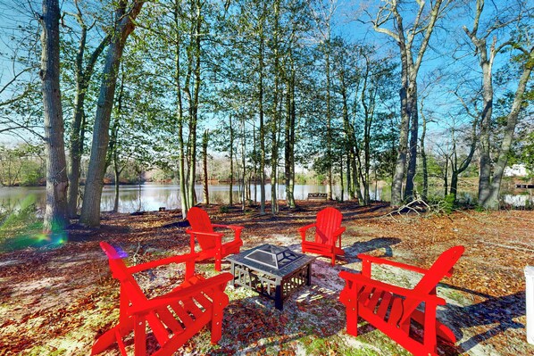 Een camping met vier rode Adirondack stoelen en een tafel, omringd door bomen en een waterpartij.