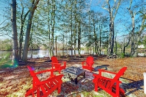 Un camping con cuatro sillas Adirondack rojas y una mesa, rodeado de árboles y una masa de agua.