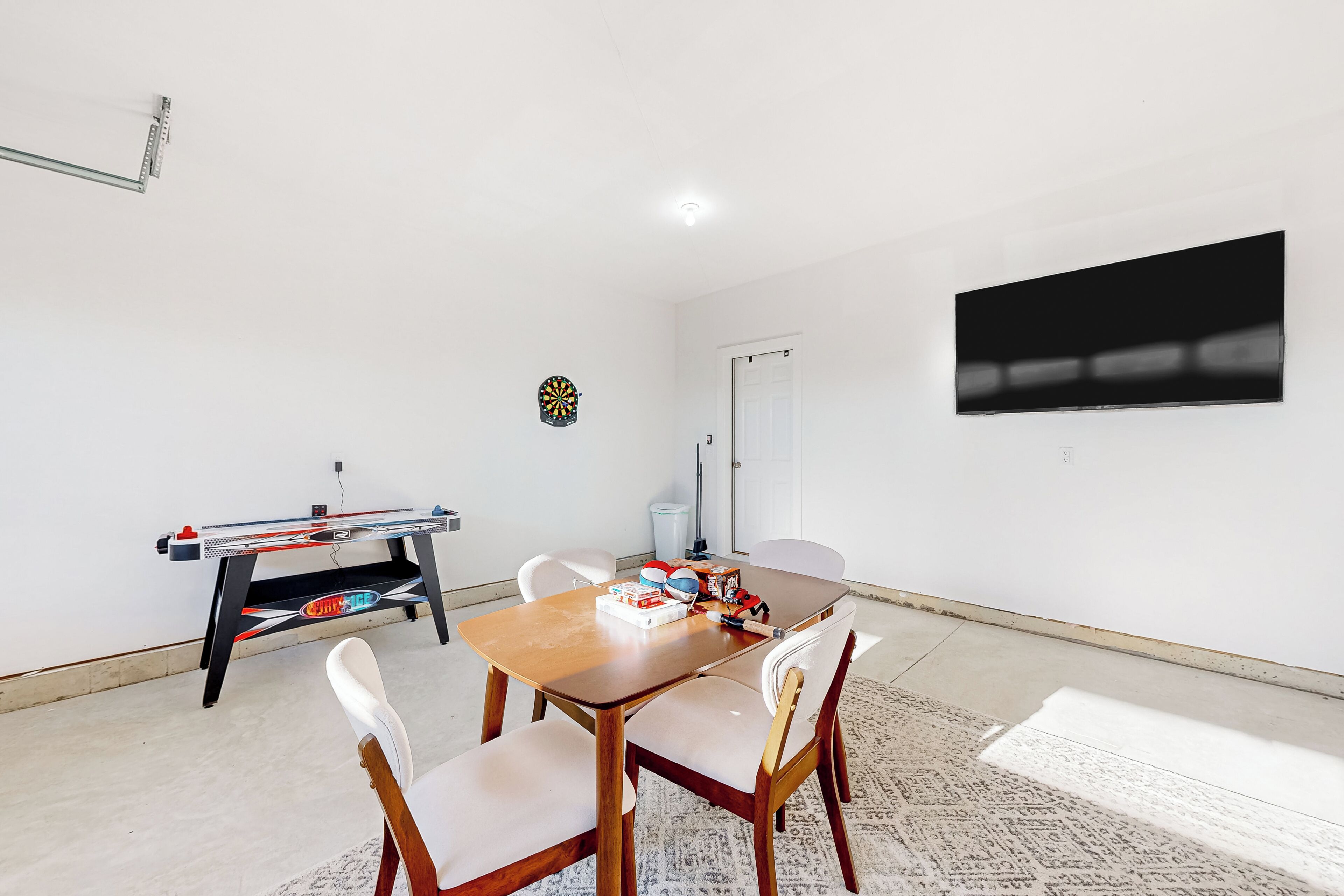 A room with a wooden table and chairs, a dartboard, a ping pong table, and a flat-screen TV mounted on the wall.