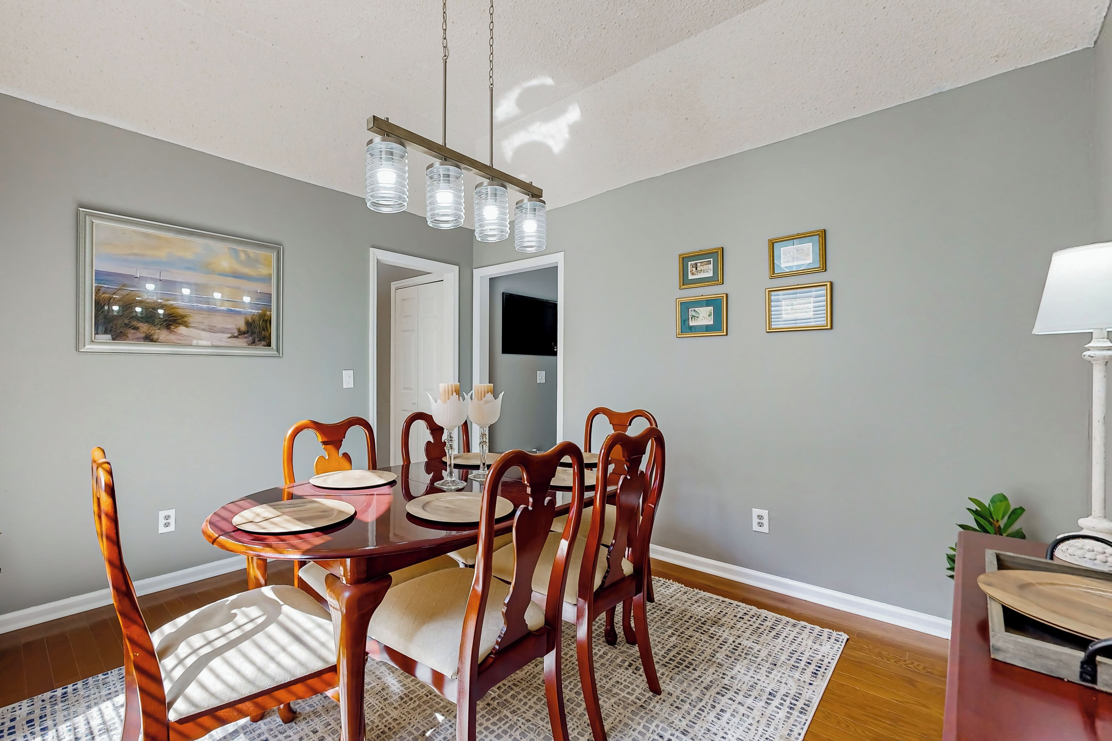 A dining area with a wooden table, chairs, and a chandelier. There are framed pictures on the wall and a plant on the side.