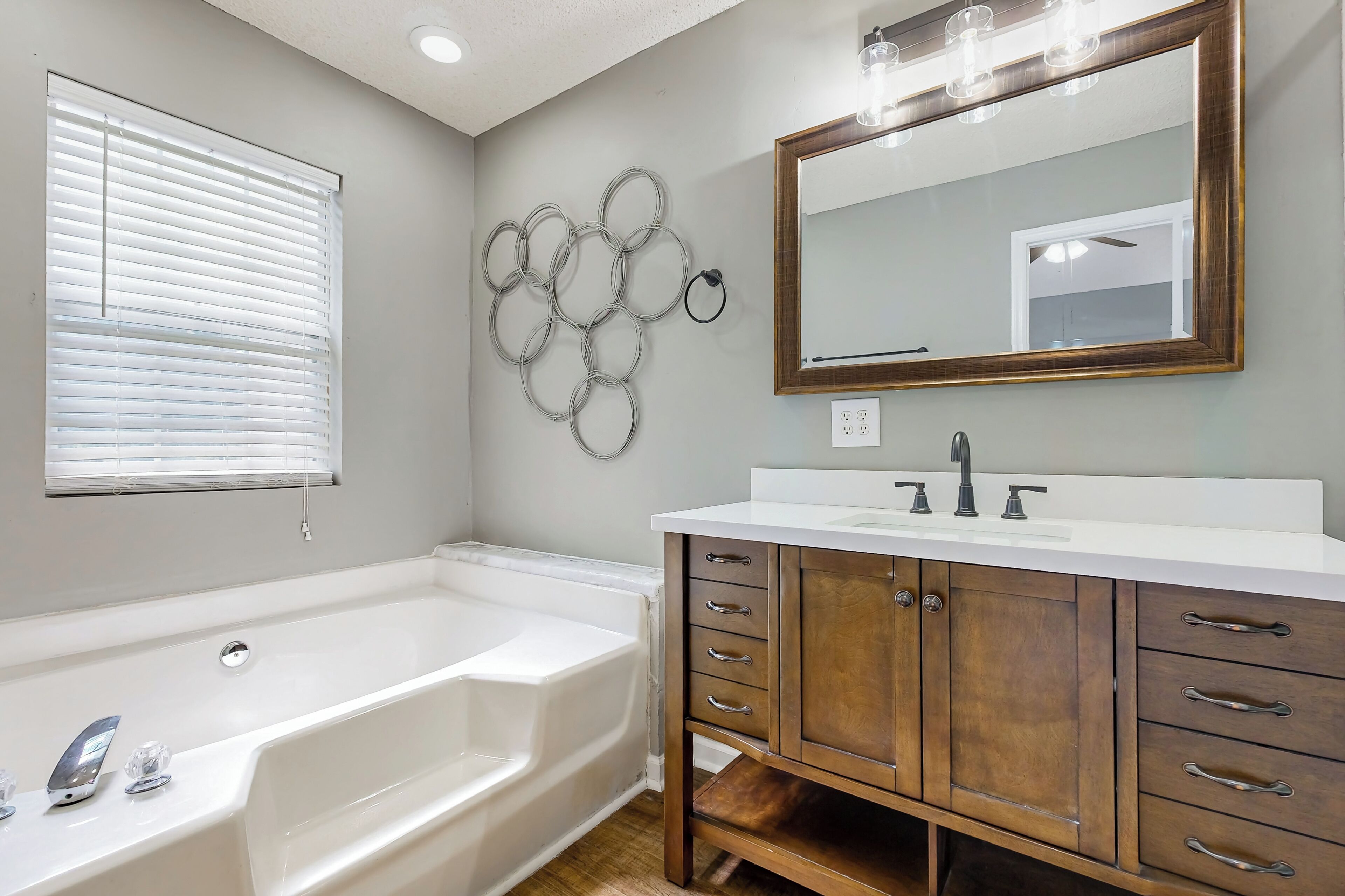 A bathroom with a white bathtub, wooden vanity with drawers, a large mirror, and a wall-mounted light fixture.