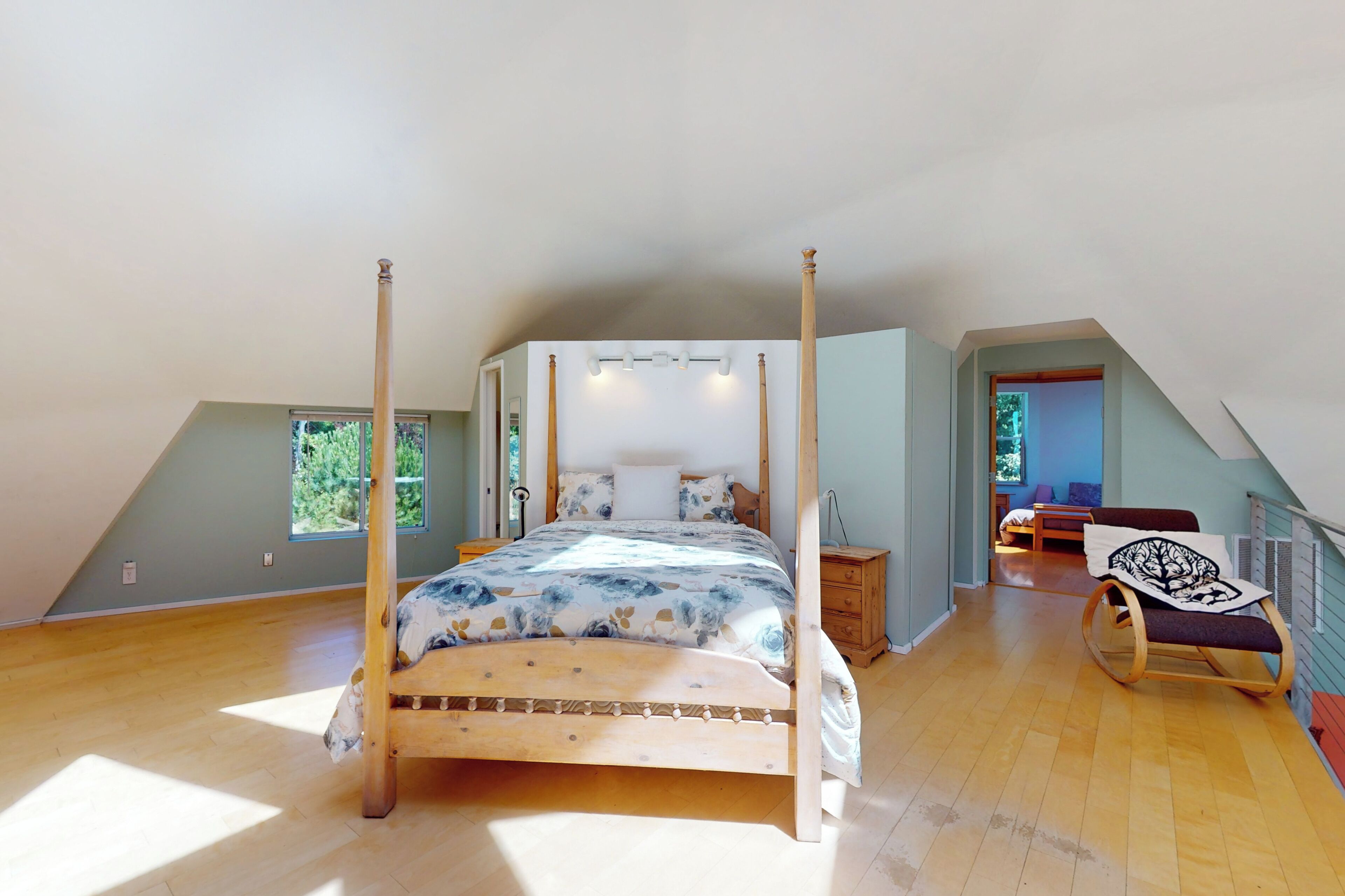 Une chambre spacieuse située sous les combles, dotée d’un lit à baldaquin, d’un plancher en bois et offrant une vue sur l’extérieur grâce à de grandes fenêtres.