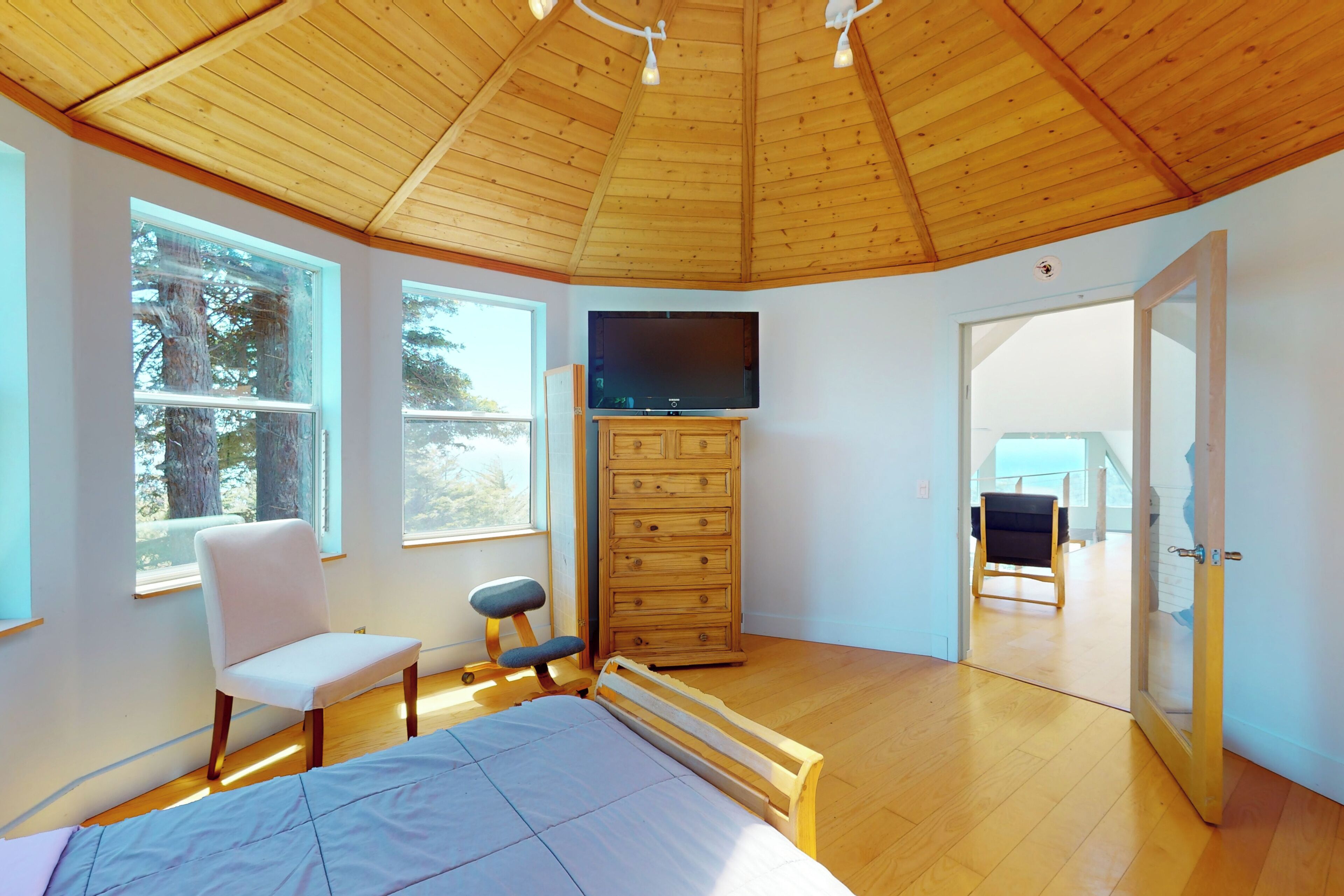 A bedroom with a wooden ceiling, a bed, a TV, a dresser, and large windows.