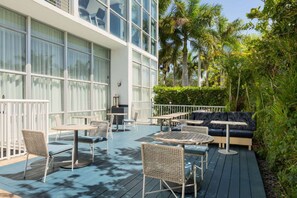 A deck with outdoor seating, including tables and chairs, surrounded by lush greenery and a building with large windows. - Waterfront Elegance Near Cruiseport | Pool & Gym (Fort Lauderdale)
