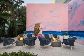 An outdoor seating area with fire pits and lounge chairs, overlooking a painted mural of a beach scene. - Waterfront Elegance Near Cruiseport | Pool & Gym (Fort Lauderdale)