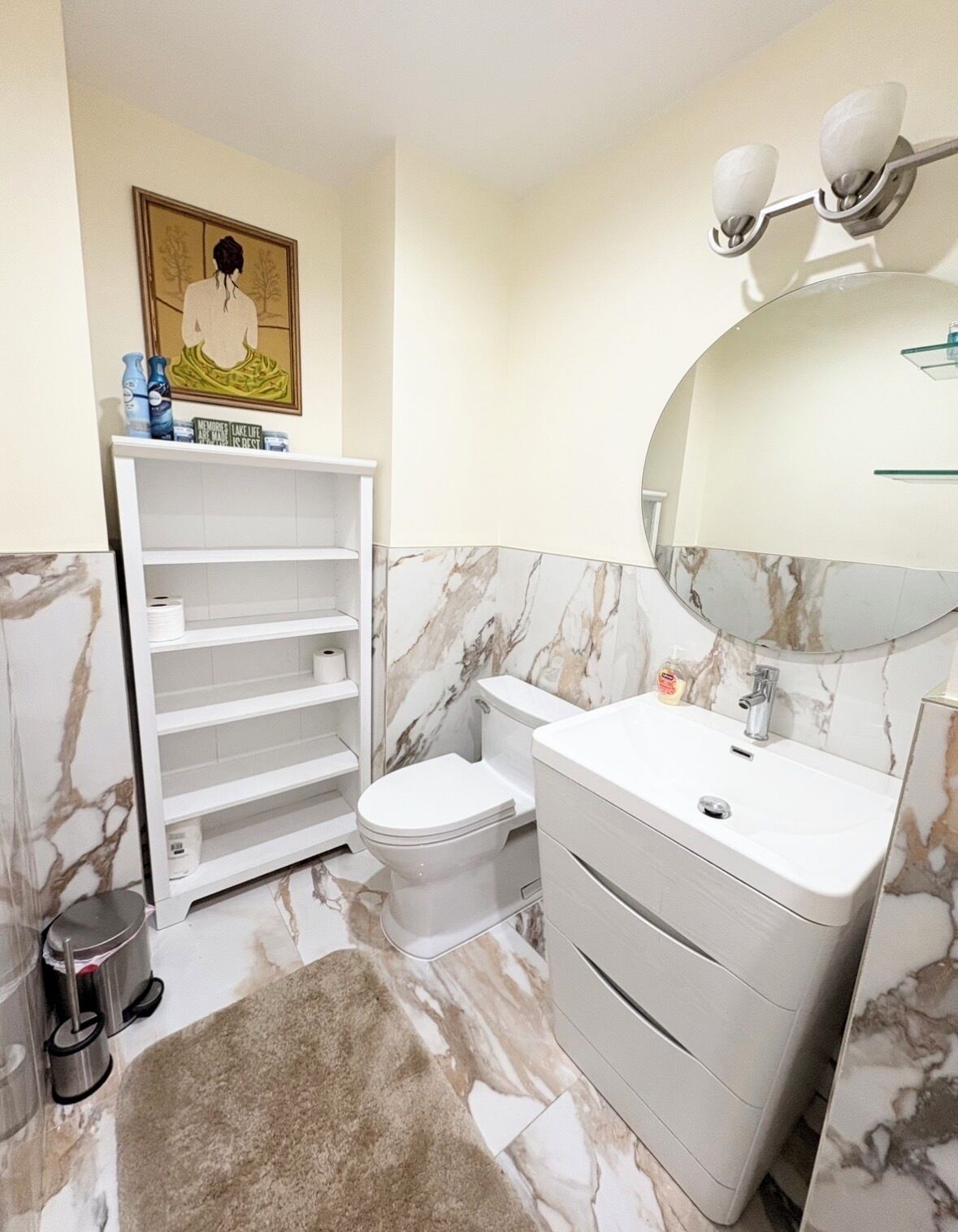 Un baño con inodoro, tocador blanco con espejo, paredes con estampado de mármol y un estante con artículos de aseo.