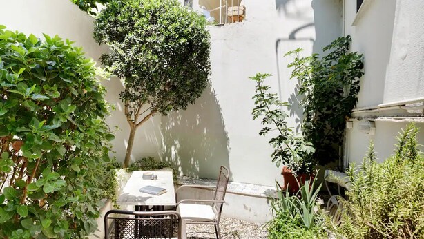 A small outdoor seating area with a table and chairs, surrounded by greenery and a white wall. - Apartment with Leafy Backyard Steps from the Acropolis (Athens)