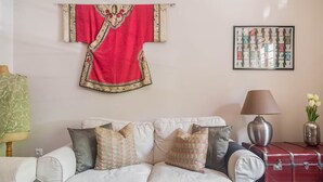 A living room with a white sofa, patterned cushions, a red decorative piece on the wall, a framed abstract artwork, a lamp, and a vintage suitcase. - Apartment with Leafy Backyard Steps from the Acropolis (Athens)