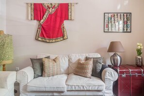 Ein Wohnzimmer mit einem weißen Sofa, gemusterten Kissen, einem roten Wanddekor, einem gerahmten abstrakten Kunstwerk, einer Lampe und einem alten Koffer.