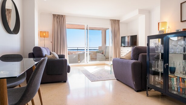 A modern living room with a glass table, two sofas, a cabinet with glassware, a TV, and a balcony with curtains. - Sea Views Lagos de Santa Maria - By Time and Home (Marbella)