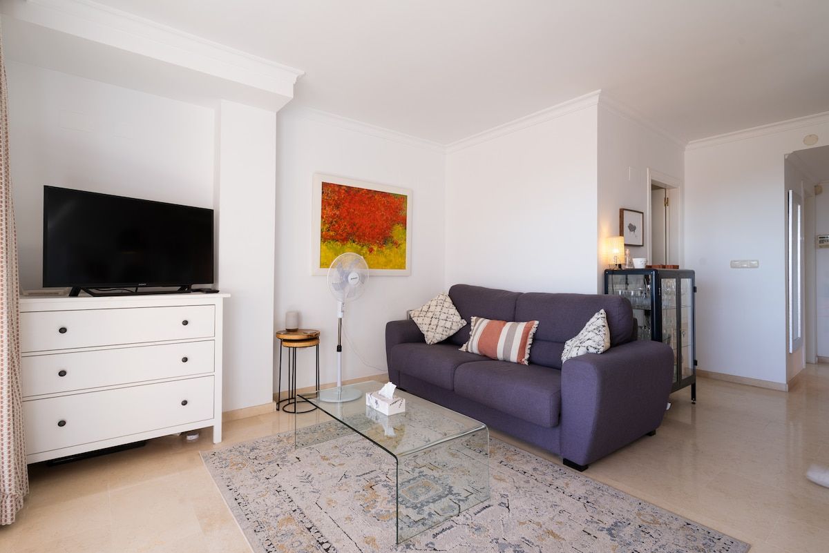 Una sala de estar con televisor de pantalla plana, sofá, mesa de centro y un cuadro en la pared.