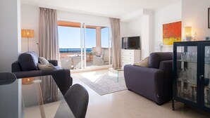 A modern living room with a glass coffee table, two sofas, a TV, and a cabinet with glassware. - Sea Views Lagos de Santa Maria - By Time and Home (Marbella)