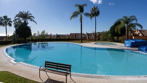 A clear blue swimming pool with a wooden bench in the foreground, surrounded by palm trees and a clear sky. - Sea Views Lagos de Santa Maria - By Time and Home (Marbella)