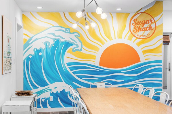 A restaurant interior with a mural of a wave and sun, a wooden table, white chairs, and a sign reading "Sugar Shack Destin, FL". - Sugar Shack*Designer*PrivatePool*BeachService (DESTIN)