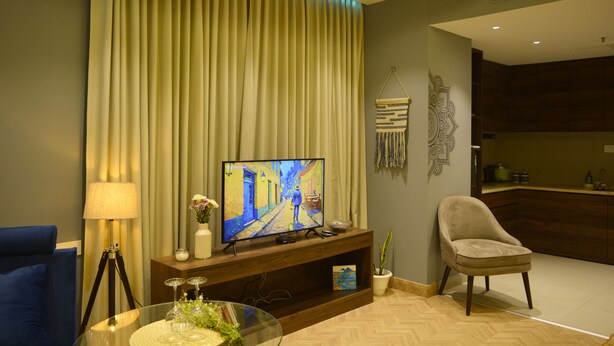 A modern living room with a flat-screen TV, a blue armchair, a glass coffee table, and a decorative wall hanging. - Luxury Skyline Studio • Near Cyberhub & Metro • NirviiHomes (Gurugram)