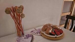 A table with a glass vase containing wooden sticks and woven balls, a wooden plate with croissants and a bowl of red jam, and a decorative heart-shaped coaster.