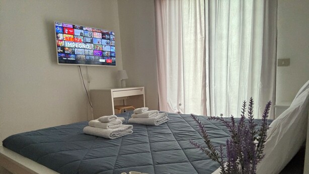 A neatly made bed with folded towels, a television displaying various channels, a bedside table with slippers, and a potted plant.