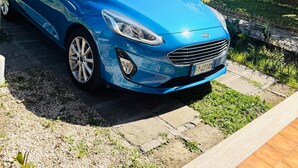 A blue car parked on a paved area with a stone wall and greenery in the background.