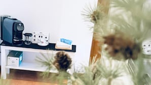 A room with a coffee machine, a small shelf with a box, and a plant in the foreground.