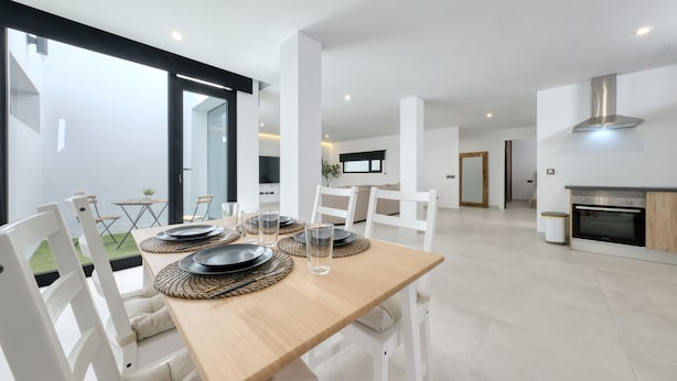 Un comedor moderno con una mesa de madera preparada para cuatro personas, sillas blancas y un vaso de agua.