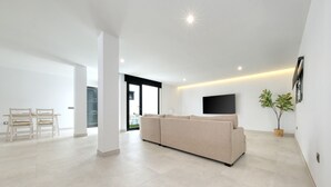 Una sala de estar moderna con un sofá color beige, un televisor de pantalla plana montado en la pared, una planta en maceta y un comedor con sillas de madera y una mesa.