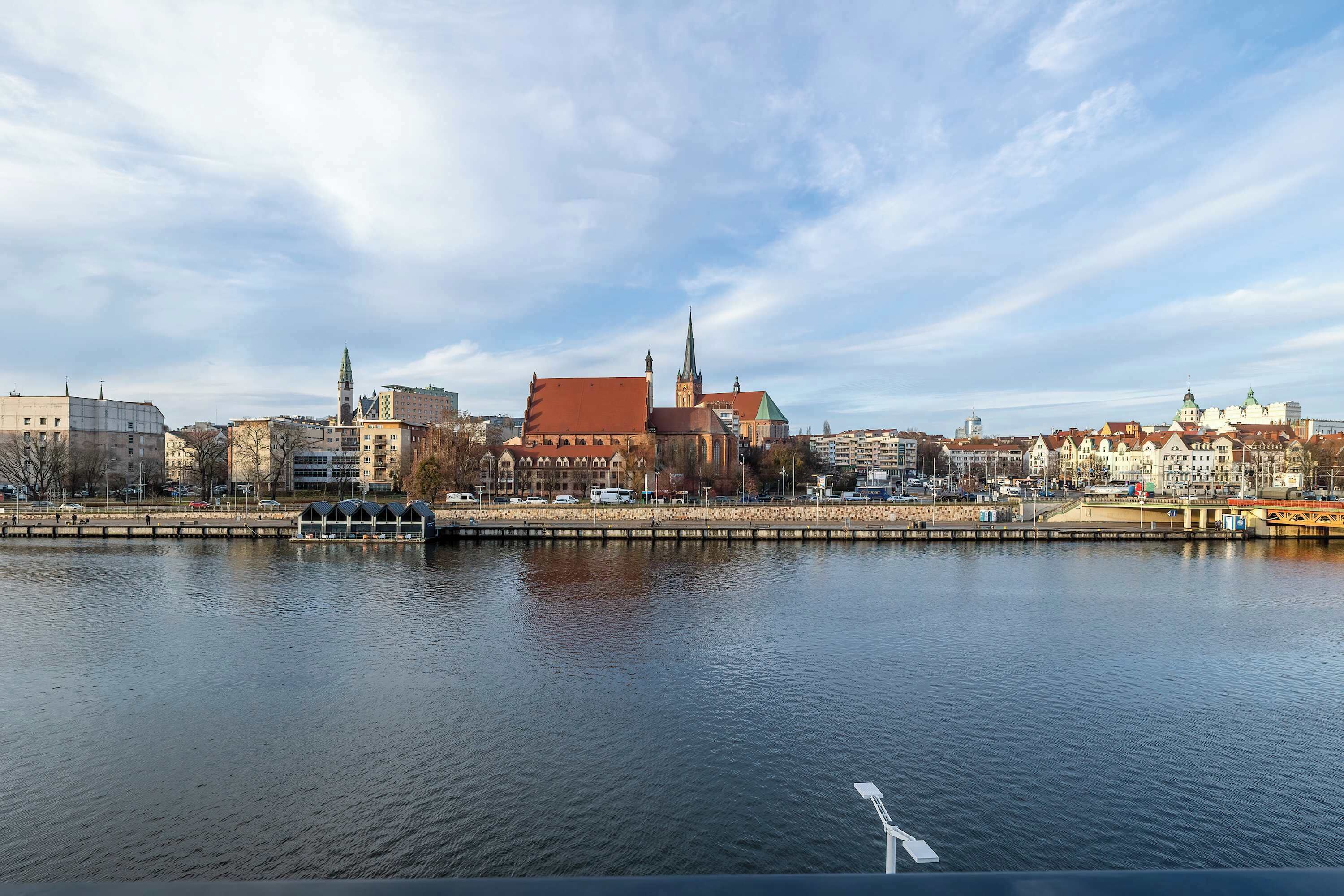 Vista de la ciudad desde la orilla de un río, con edificios históricos, incluyendo una iglesia destacada con una aguja alta.