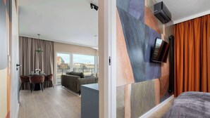 Een moderne hotelkamer met een groot bed, een aan de muur bevestigde televisie, een zithoek met een tafel en stoelen, en uitzicht op de stad door grote ramen.