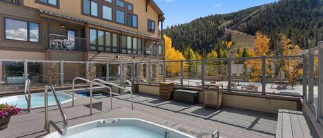 A rooftop pool area with a hot tub, outdoor seating, and a view of a forested mountain.
