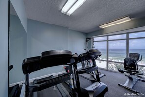Un gimnàs amb cintes de córrer, una bicicleta estatica i una màquina d'a rem. Té grans finestres amb vista a la platja.