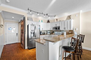 A kitchen with white cabinets, stainless steel appliances, a wooden floor, and a bar area with stools. - NEW Emerald Isle 405, FREE Beach Service, Heated Pool, Hot Tub, Grill & Perks (Panama City Beach)