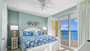 A bedroom with a bed, a nightstand, a ceiling fan, and a view of the ocean through a sliding glass door. - NEW Emerald Isle 1308, FREE Beach Service, Heated Pool, Hot Tub, Grill &Perks (Panama City Beach)