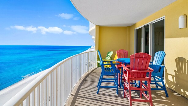 A balcony with a view of the ocean, featuring a table and colorful Adirondack chairs. - NEW Emerald Isle 1308, FREE Beach Service, Heated Pool, Hot Tub, Grill &Perks (Panama City Beach)