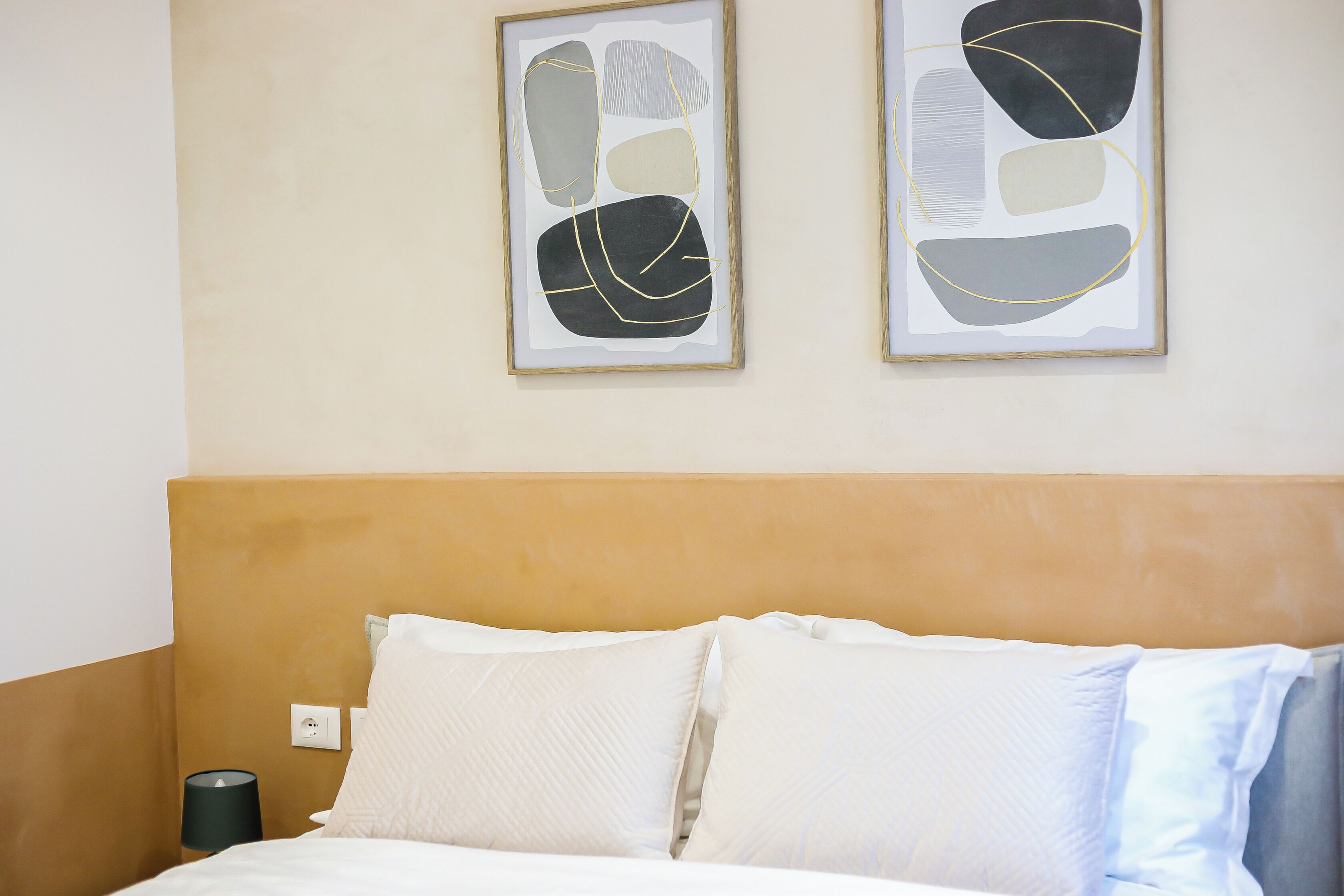 Een net opgemaakt bed met witte lakens en twee kussens, een bedlamp en abstracte kunst aan de muur.