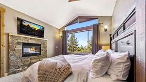 A bedroom with a stone fireplace, a large bed, a television, and a view of trees through a window. - Heavenly Luxe Chalet - Hot Tub, Theatre, Game Room (South Lake Tahoe)